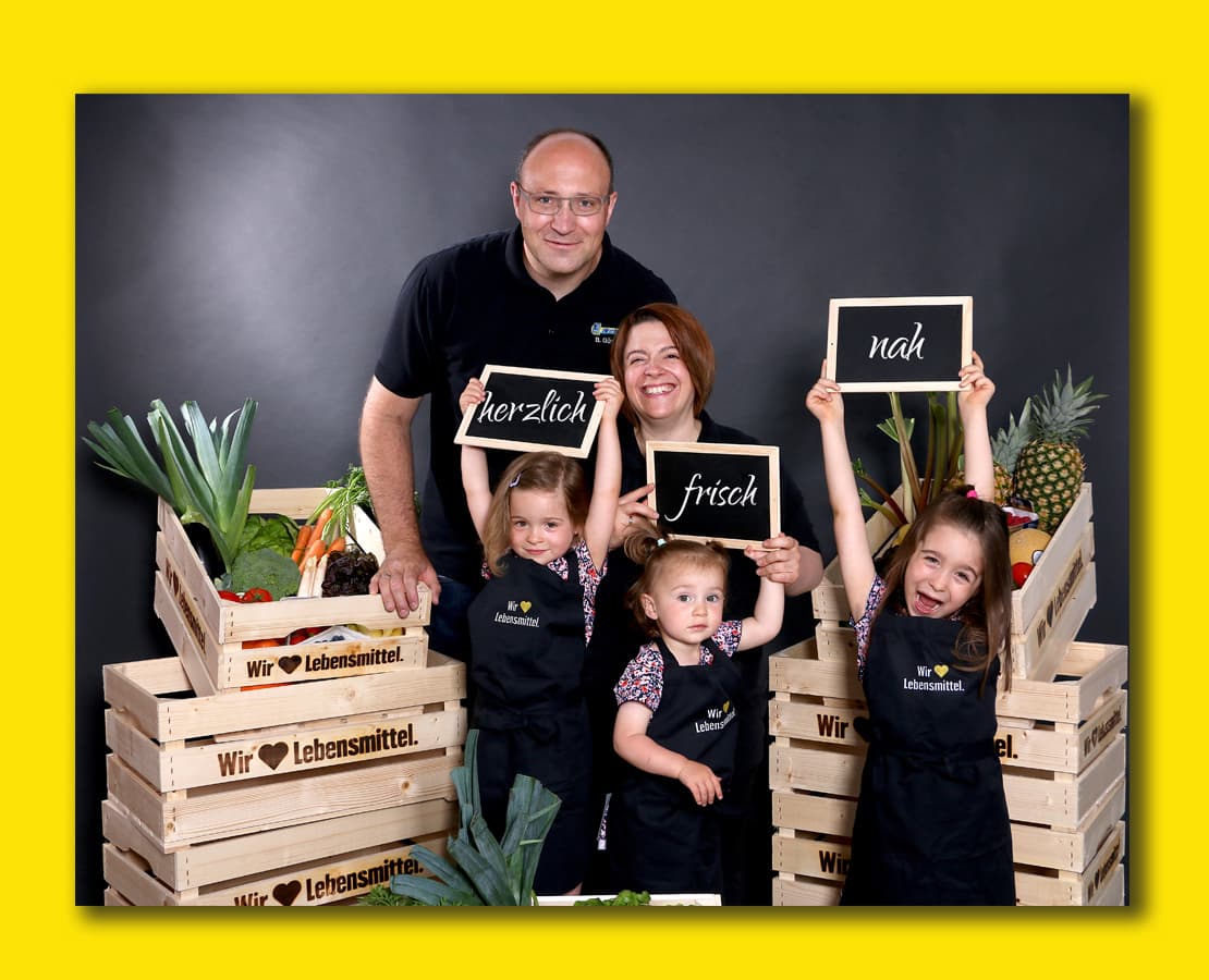 Ein fröhliches Foto der Familie Weinle zwischen Obst- und Gemüsekisten auf gelbem Hintergrund