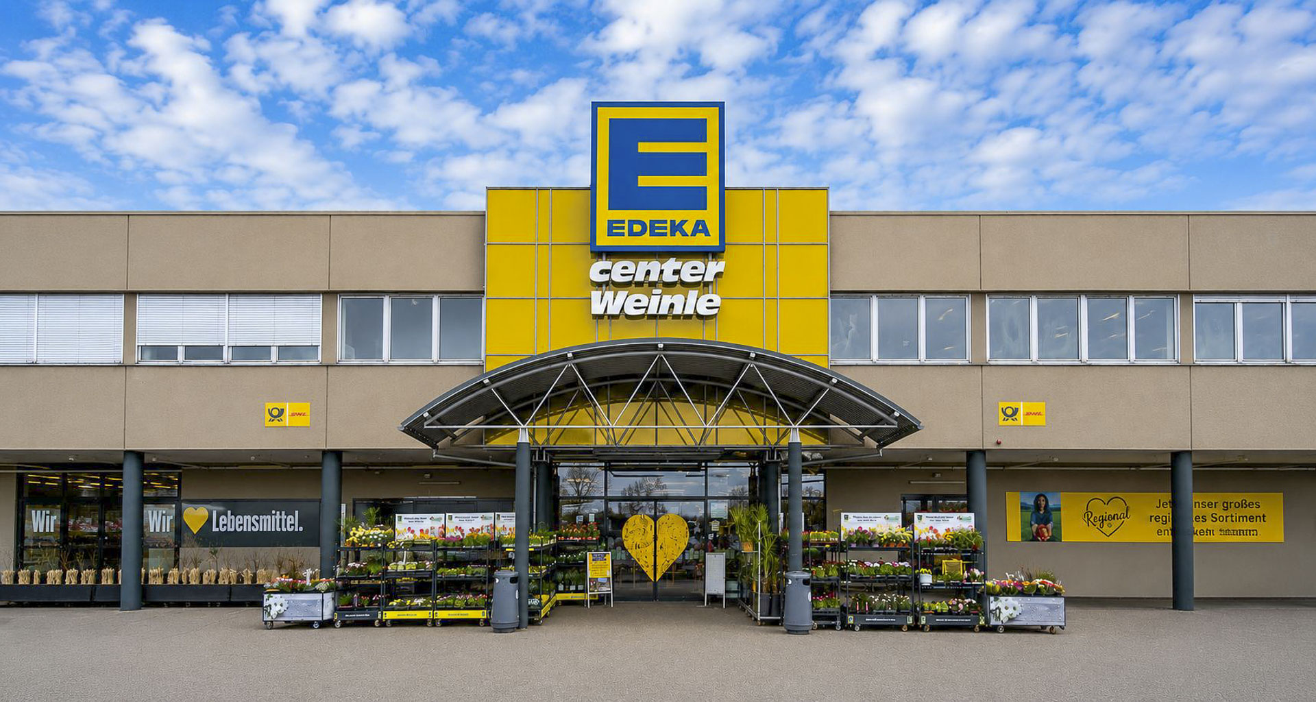 Frontansicht des modernisierten EDEKA Center Weinle mit symmetrischer Architektur, großem Eingangsbereich und Blumenverkauf vor dem Markt unter bewölktem Himmel.